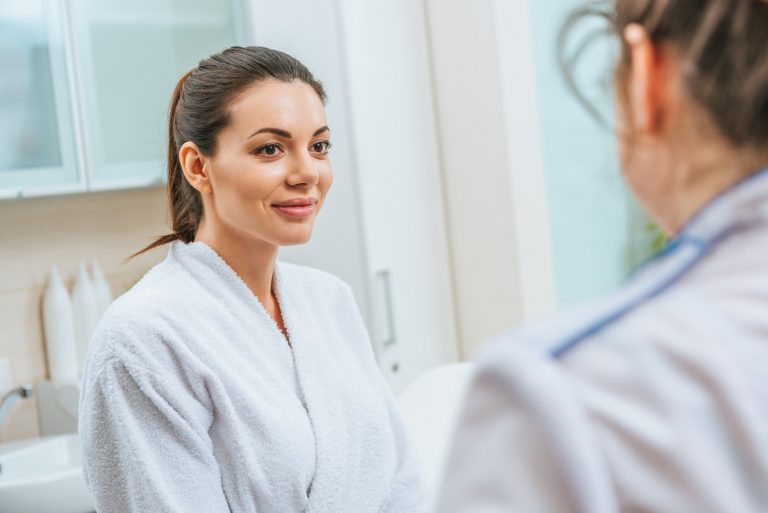 Beautiful smiling young woman in white bathrobe looking at cosmetologist in beauty clinic
